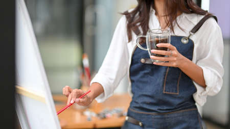 Cropped shot, female artist holding coffee while drawing on canvas easel, relaxed hobby, art work, art therapyの写真素材