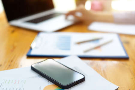 Closeup smartphone on working desk with property investment report, blurred female hand working on...