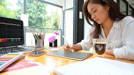 Young female graphic designer sitting and using stylus and tablet sketching new art model at studio, creative occupationの写真素材