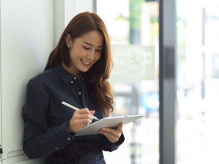 Gorgeous business female leaning on wall and working in digital tablet at office spaceの写真素材