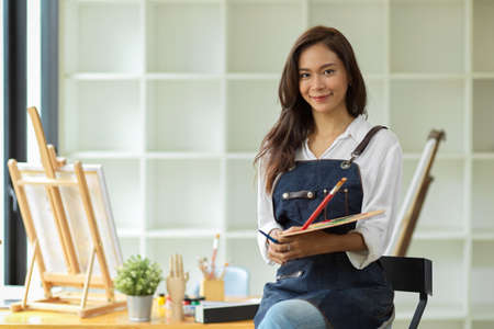 Elegant asian artist woman sitting and holding colour palette and paintbrush with blurred art studio in the backgroundの写真素材