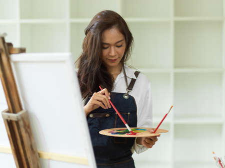 Hobby and leisure concept, Young asian female art student practicing oil colour paint on canvas easel at art school, holding colour paletteの写真素材