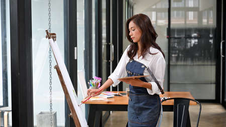 Attractive female artist paint picture on large canvas easel with oil colour, holding palette. art studio or art workshopの写真素材