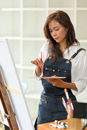 Portrait young attractive female artist painting on canvas easel, mix different colour on palette at art studioの写真素材