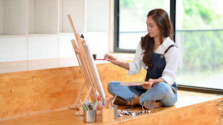 Pretty female artist sitting on the floor, painting on canvas easel with painting supplies, joining in art workshopの写真素材