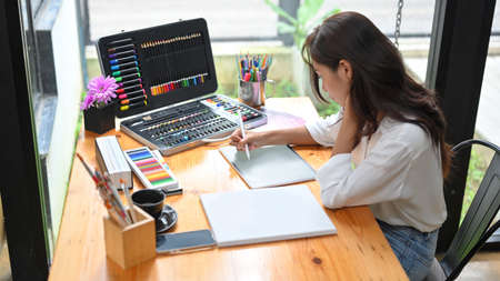 Female artist sketching picture layout on digital tablet before draw on canvas poster at studio, colouring supplies on wooden tableの写真素材