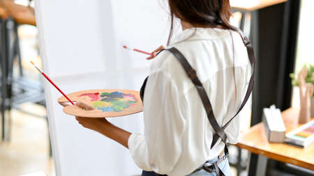 Female artist standing backwards looking on canvas, holding colour palette and paintbrush at art studioの写真素材