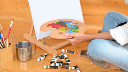 Closeup, female art student sitting and painting on canvas, holding palette, colour tubes on floor, painting toolsの写真素材