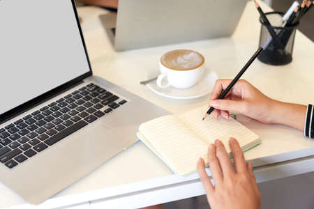 Cropped image of woman take notes on notebook next to laptop computer mockup and coffee mug at officeの写真素材