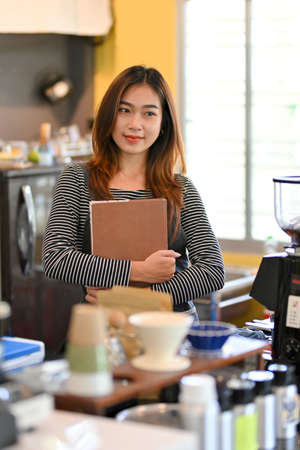 Portrait of cheerful female barista smiling, looking away, standing near coffee machine at coffee shopの写真素材