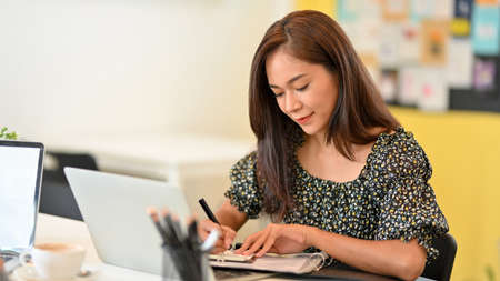 Attractive business assistant taking notes in personal planner book, working in front of laptop computer at officeの写真素材
