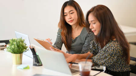 One of business team taking notes on paper clipboard while her colleague searching idea from website via laptop at officeの写真素材
