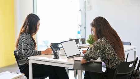 Two of young hardworking female working at office with electronic devices, using laptop, office supplies on deskの写真素材