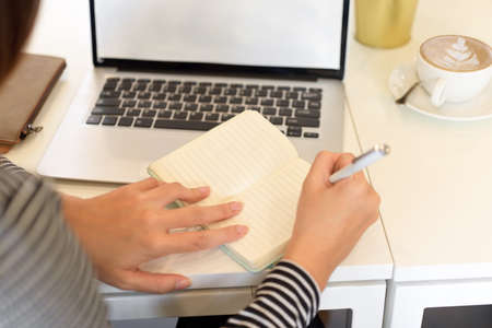 Closeup, Female accountant taking notes about company's tax on notebook with opened laptop on desk, laptop screen mockupの写真素材