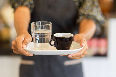 Cropped image of black coffee mug with a glass of water on serving tray hold by female barista at...