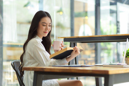 Side view, Attractive woman enjoy reading book and drinking coffee at coffee shop, relaxing time, leisure activityの写真素材