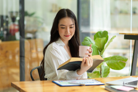 Young adult asian businesswoman drinking coffee and reading novel for take a break from work, sitting at coffee shop, relaxing timeの写真素材