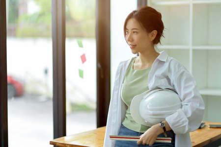 Young female engineer looking out of window, holding ruler and hardhat in engineers officeの写真素材