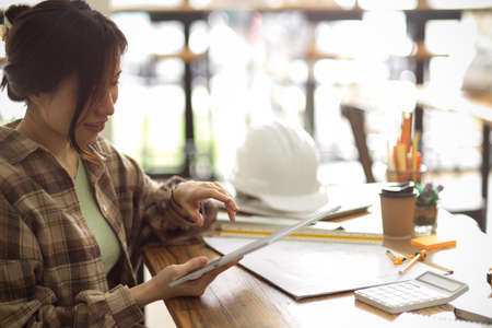 Young female architecture looking building scale and plan on digital tablet at engineering office room, architectural projectの写真素材