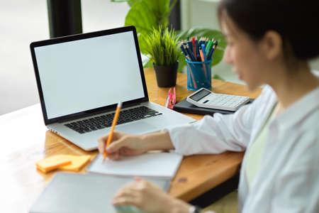 Female university student online learning, using tablet and laptop for study, taking notes or lecture on notepad with pencil in dormitory co-working spaceの写真素材