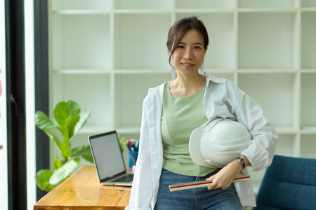 Portrait of young female architect with hardhat and ruler leaning on working desk in modern engineers officeの写真素材