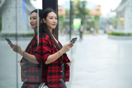Beautiful young woman using an application in her smartphone outside of shopping mall, looking away,...