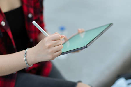 Closeup female's hands using portable tablet with stylus pen, sitting in park, blurred backgroundの写真素材