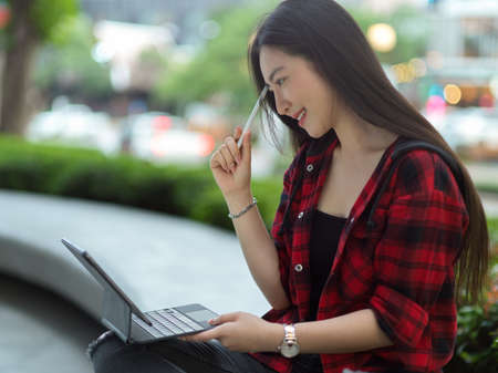 Attractive asian woman sitting in park, thinking about work, finding ideas, working on portable tablet outdoorsの写真素材