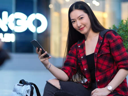 Beautiful woman in red sweater with smartphone and backpack sitting in front of shopping centre, girl posing with digital deviceの写真素材