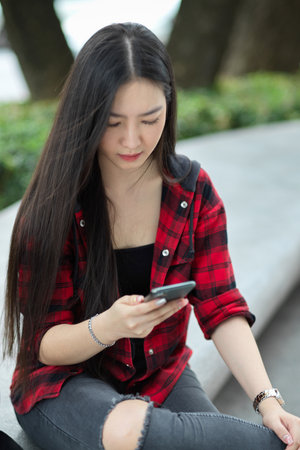 Female teenager sitting in city street, messaging to her friends, using smartphone for wasting time, scrolling on social mediaの写真素材
