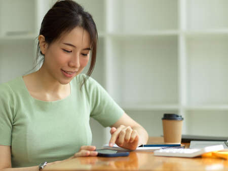 Pretty young woman touching screen, using smartphone on worktable in office, texting, messaging, playing mobile game, online shopping, business contact, reading emailの写真素材