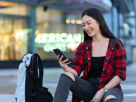 Joyful of asian young female traveller watching funny video on phone, chatting with friends, using smartphone, waiting friends in front of shopping store in eveningの写真素材