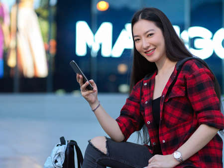 Attractive hipster woman sitting in front of city shopping mall and using smartphone, blurred shopping banner in backgroundの写真素材