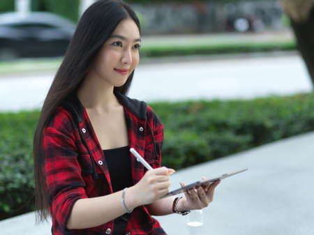Young attractive and beautiful asian woman sitting in city park square using tablet with stylus pen...