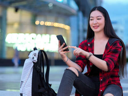 Solo female backpacker using mobile phone to booking online taxi cab service and booking hotel, sitting in city street in eveningの写真素材
