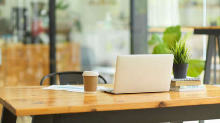 Image of Laptop computer stand on wooden table with coffee cup, report, books, indoor plant in coffee shop co-working spaceの写真素材