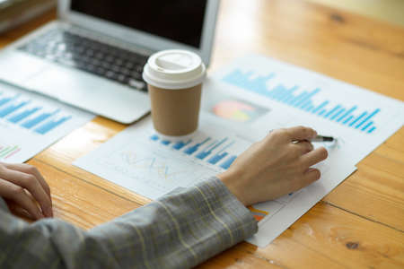 Closeup hand, businesswoman planning and analysing business sale performance from business data report. laptop and take away coffee cup on table, pen pointingの写真素材