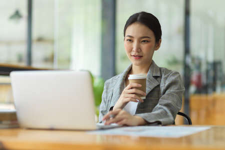 Success businesswoman working in office, looking on laptop computer screen, thinking and analysing business strategy while holding coffee cup, relax workerの写真素材