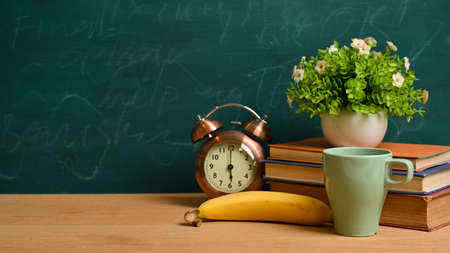Back to school concept. Books, vintage alarm clock, banana, mug, plant and mockup space on wooden tabletop over green blackboardの写真素材