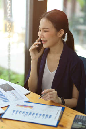 Confident young asian businesswomen talking on mobile phone with business partners while sitting at her working place in office, financial report papers on deskの写真素材