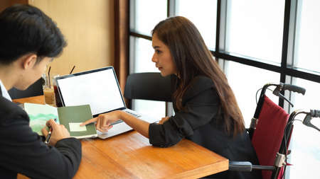 Attractive businesswoman in wheelchair explaining work to her co-worker in modern office. laptop computer mockupの写真素材