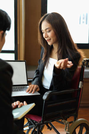 Portrait of asian beautiful businesswoman in wheelchair presenting her work on laptop to manager in officeの写真素材