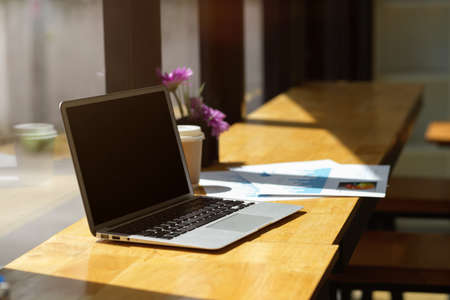 Opened laptop computer black screen mockup and paperworks on wooden table with sunlight through window in coffee shopの写真素材