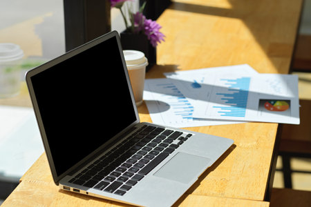 Generic laptop computer with black screen and report papers on wooden table near the windowの写真素材