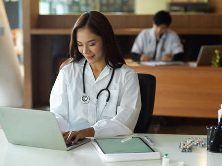 Asian young female doctor in white gown with stethoscope working on laptop computer in officeの写真素材