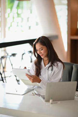 Happy young female physician looking at symptoms diagnosis papers and taking notes on medical clipboard at her deskの写真素材