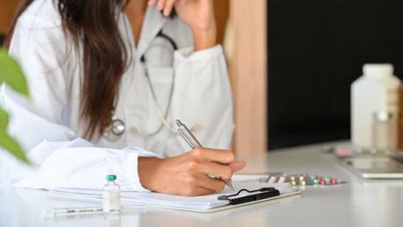 Cropped shot of female practitioner or pharmacist writing a medical prescription on medicine record clipboard. vaccine vial, pills, vitamin bottles on deskの写真素材