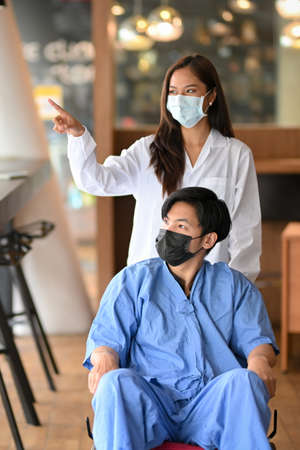Doctor or caregiver with disabled male patient in wheelchair wearing protective masks at hospital. Female doctor encouraging handicapped patientの写真素材