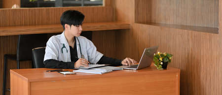 An Asian doctor sits at his desk works on laptop computer, examining the patient's medical history in preparation for an examination and diagnosis.の写真素材