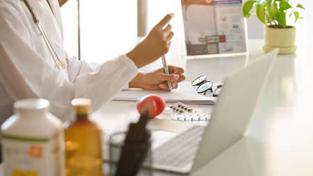 Pharmacist is researching medical drugs and writing medical prescriptions. On the work table, there are pills, vitamin bottles, a laptop, and glasses. Cropped imageの写真素材